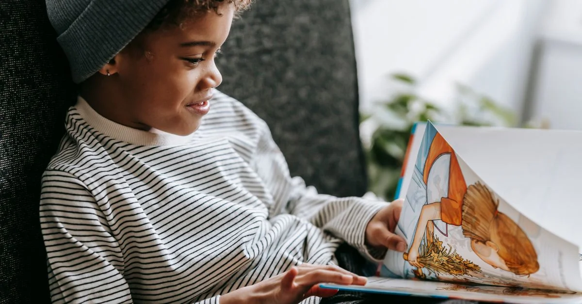 Un garçon heureux avec des cheveux bouclés profite de la lecture d'un livre tout en se relaxant à l'intérieur, entouré de lumière naturelle.