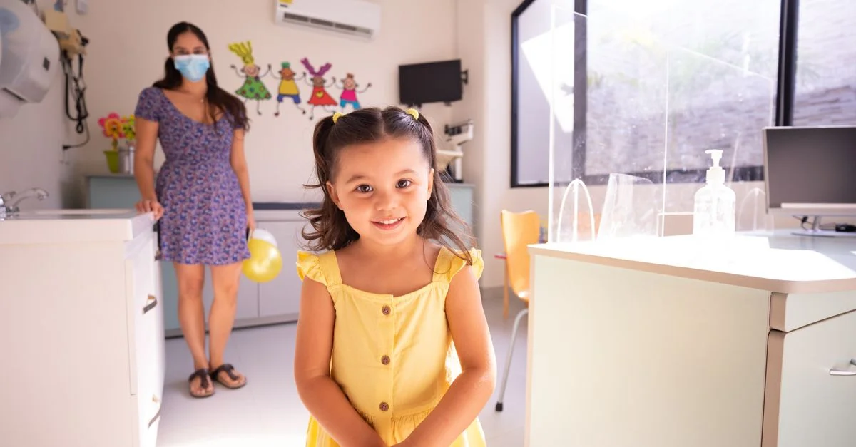 Une enfant joyeuse en robe jaune dans une clinique pédiatrique avec un médecin en arrière-plan.