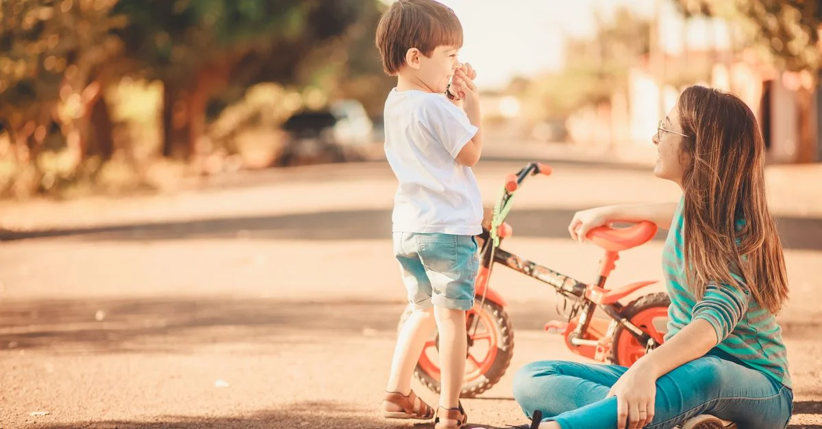 Une jeune femme est assise avec son enfant dans une rue de la ville, avec un tricycle à proximité dans un cadre ensoleillé et chaleureux.
